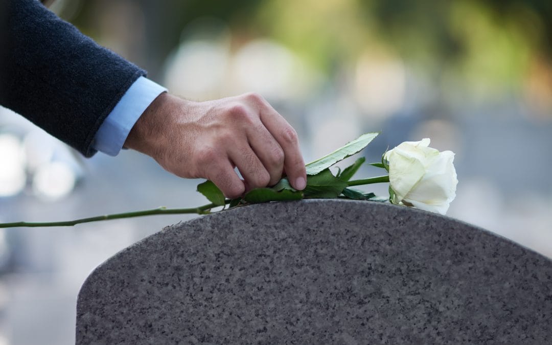 A hand places a white rose on a gray tombstone during graveside services in a cemetery.